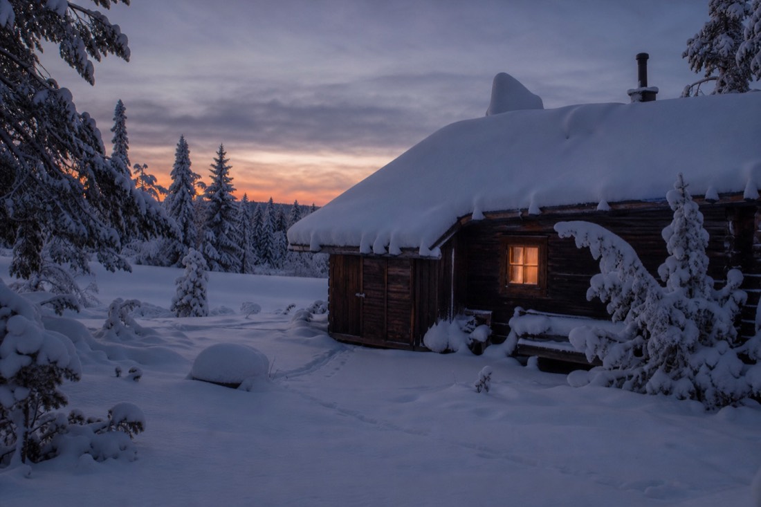A wooden Lapland cabin under thick snow at dusk, one window lit warm